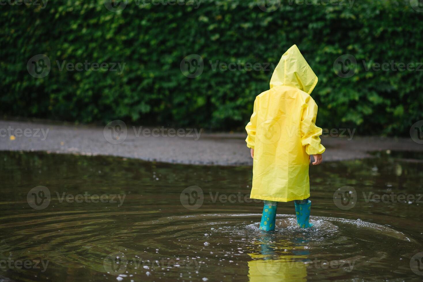 peu garçon en jouant dans pluvieux été parc. enfant avec parapluie, imperméable manteau et bottes sauter dans flaque et boue dans le pluie. enfant en marchant dans été pluie Extérieur amusement par tout temps. content enfance photo