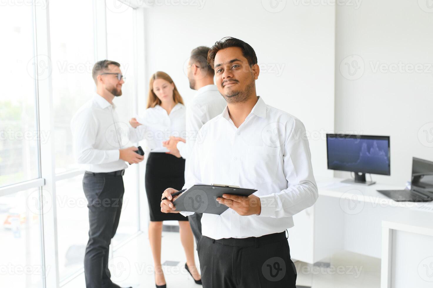 portrait de souriant Indien homme d'affaire permanent dans moderne Bureau sur collègues Contexte photo