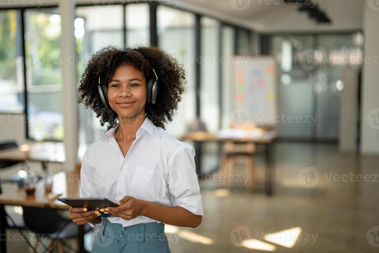 africain américain Université étudiant avec écouteurs dans main avec tablette permanent souriant à la recherche vers l'avant dans café ou espace de travail photo