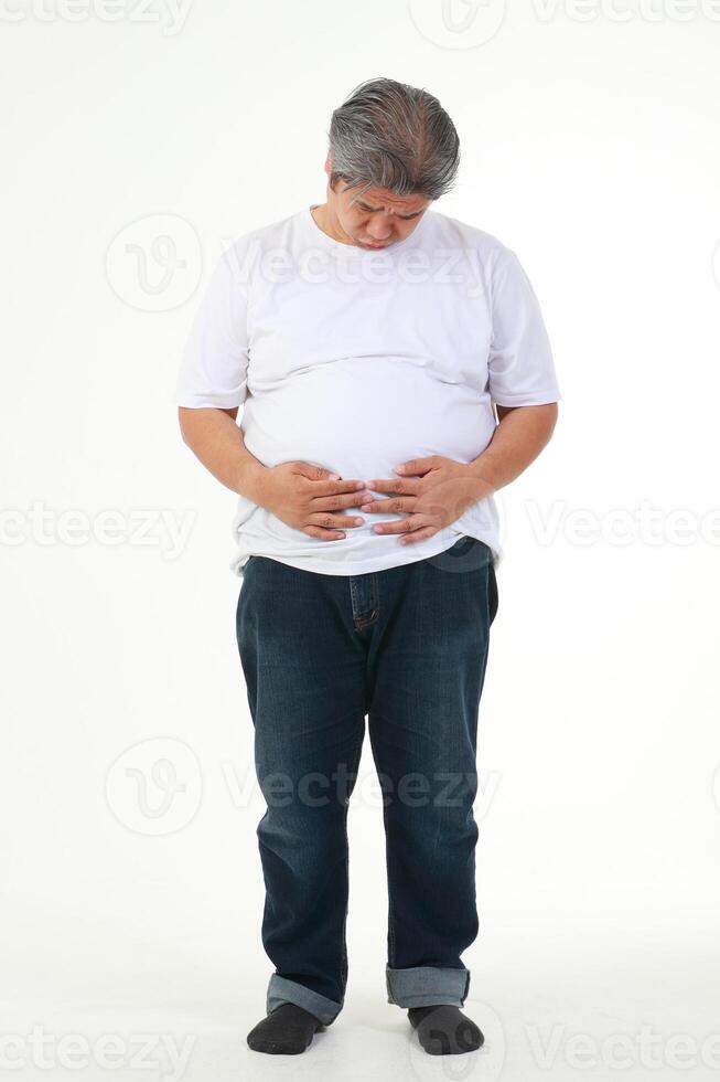 une graisse asiatique homme dans une blanc T-shirt se tenait en portant le sien ventre. il se sentait inconfortable, abdominal distension, et avait symptômes de maladie. poids perte concept. santé se soucier. blanc Contexte photo