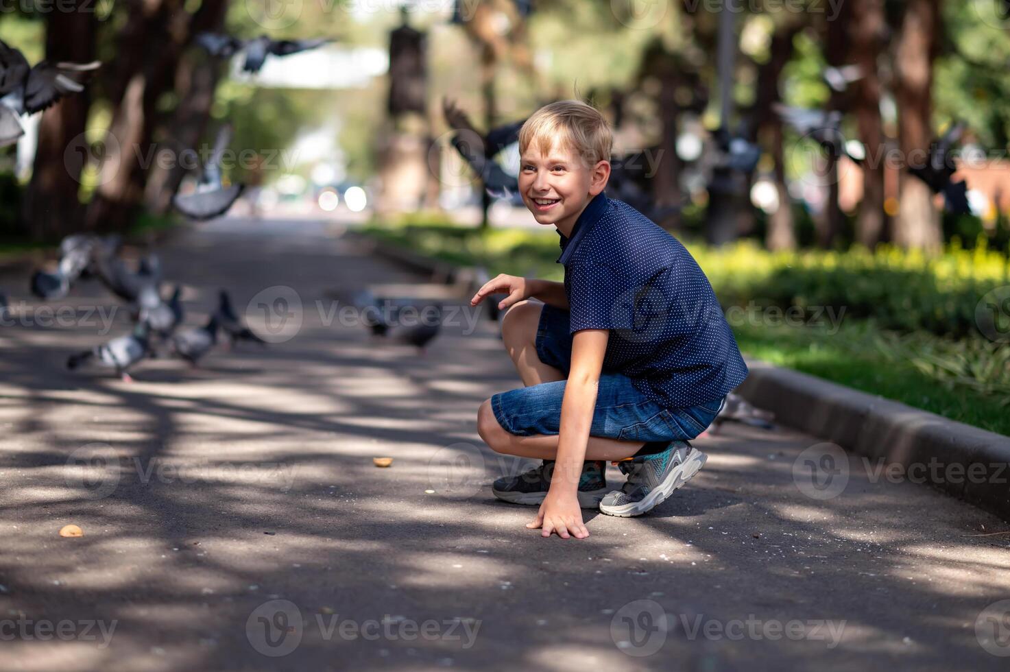 une garçon dans une bleu T-shirt est assis sur le asphalte dans le parc et regards à le pigeons. photo