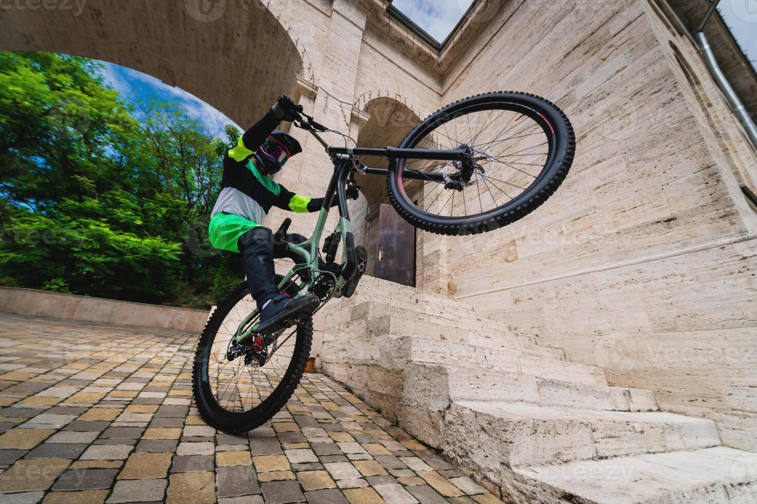 homme permanent sur un roue de une Montagne vélo. horizontal coup en plein air dans le ville avec architecture dans le arrière-plan, large angle. gars effectue une des sports tour photo