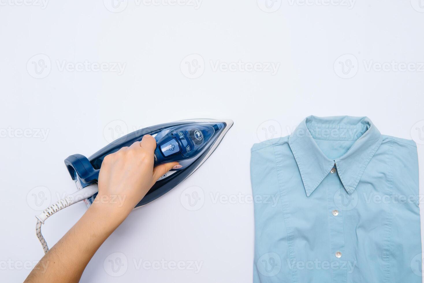 femelle main repassage vêtements Haut vue isolé sur blanc Contexte. Jeune femme avec le fer repassage homme chemise vu de au dessus pendant travaux ménagers. bleu le fer sur blanc table photo