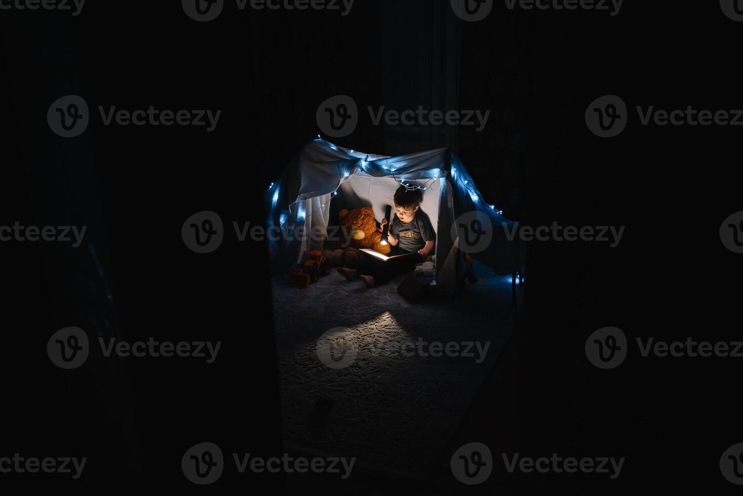 enfant garçon en train de lire avec livre et lampe de poche et nounours ours dans tente. avant Aller à lit. photo