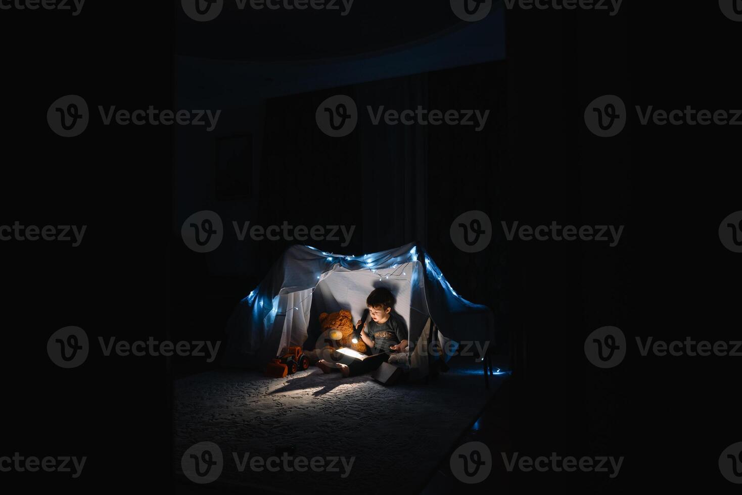 enfant garçon en train de lire avec livre et lampe de poche et nounours ours dans tente. avant Aller à lit. photo