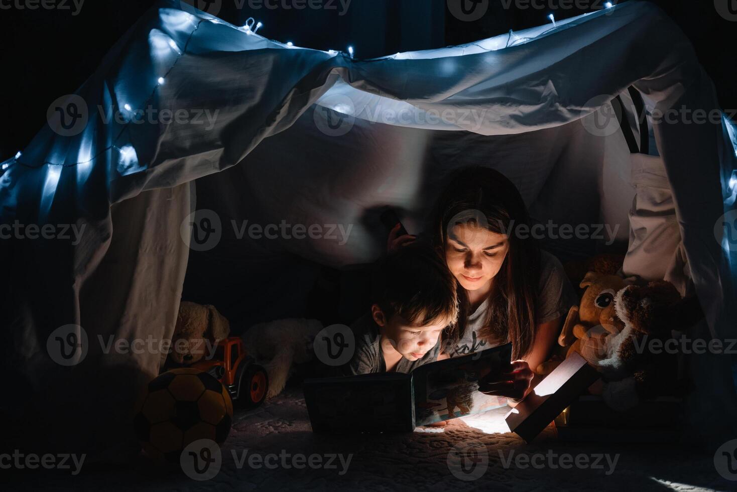 content famille mère et enfant fils en train de lire une livre avec une lampe de poche dans une tente à maison. famille concept. photo