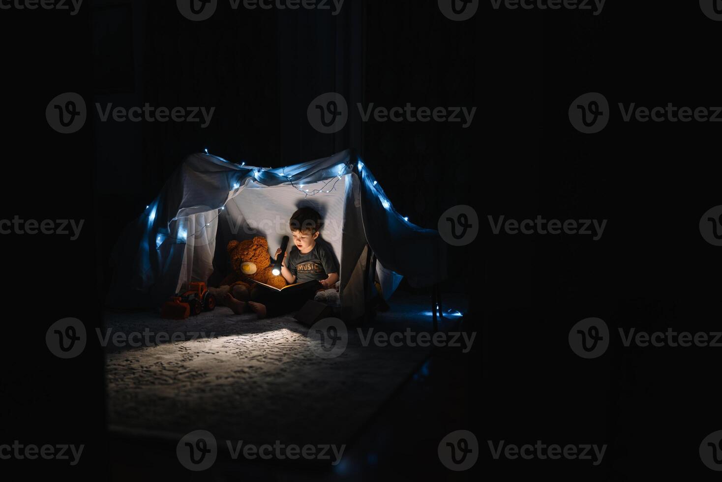 enfant garçon en train de lire avec livre et lampe de poche et nounours ours dans tente. avant Aller à lit. photo