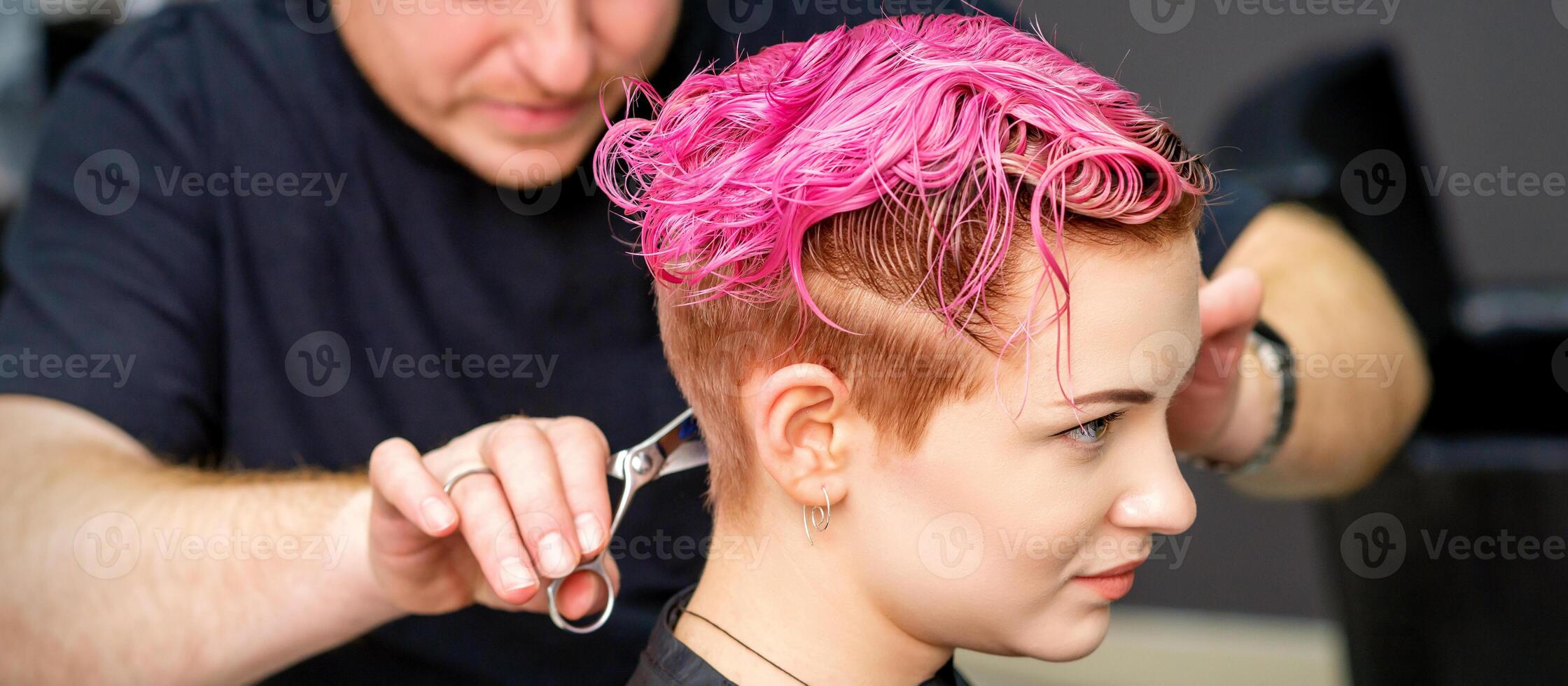 femme ayant une Nouveau la Coupe de cheveux. une Masculin coiffeur est Coupe teint rose court cheveux avec les ciseaux dans une cheveux salon. photo