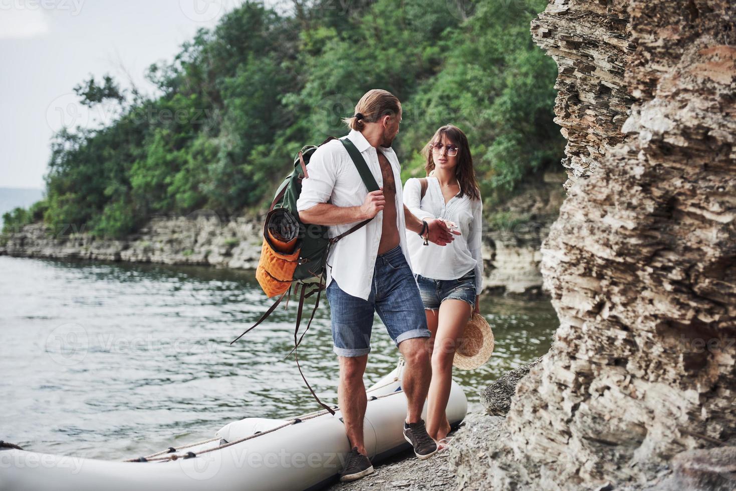 mignon jeune et couple sur fond de rivière. un gars et une fille avec des sacs à dos voyagent en bateau. concept d'été de voyageur photo