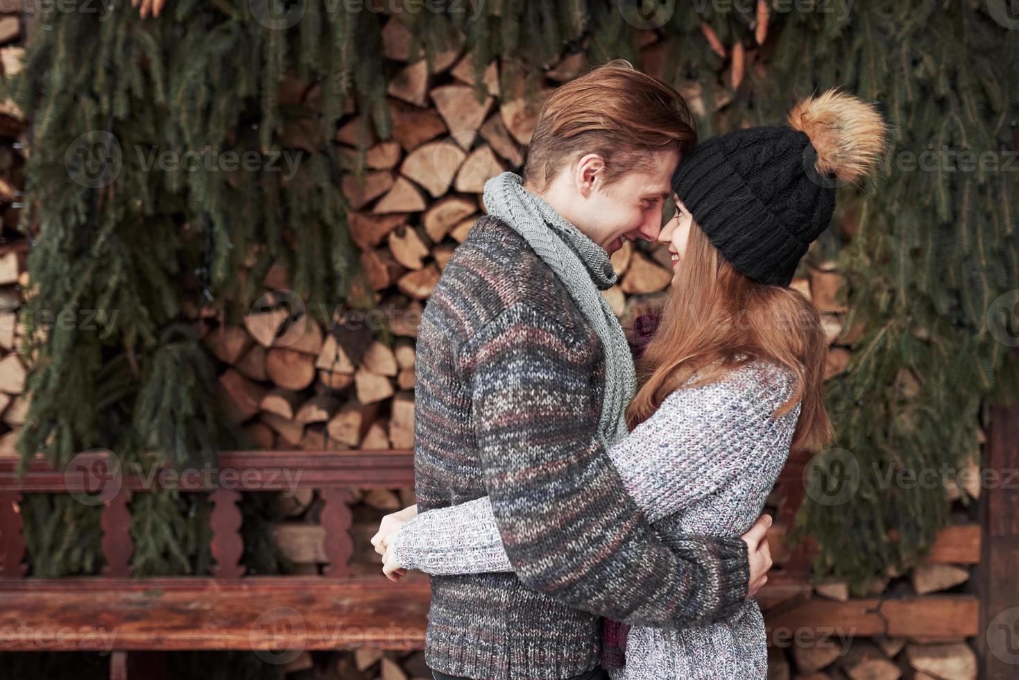 concept de noël et de couple - homme et femme souriants en chapeaux et écharpe étreignant sur une maison de campagne en bois et fond de neige photo