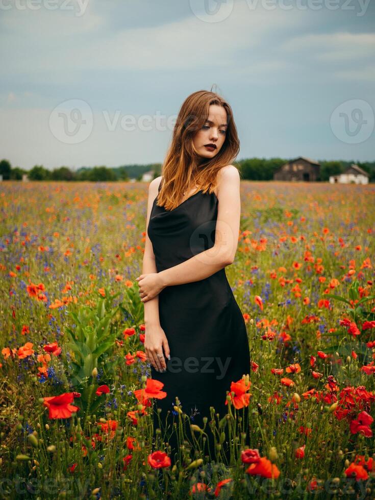 magnifique Jeune fille dans une noir soir robe posant contre une coquelicot champ sur une nuageux été journée. portrait de une femelle modèle en plein air. pluvieux temps. gris des nuages. photo