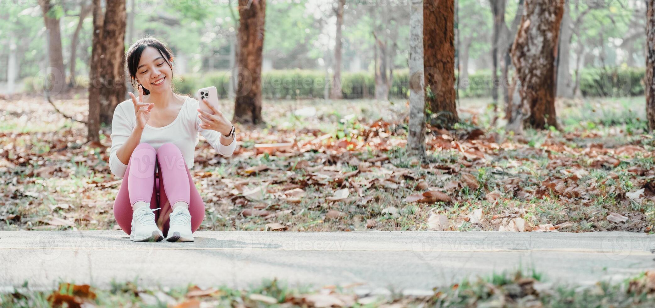 femme assise sur une parc chemin fait du une paix signe tandis que prise une selfie, avec l'automne feuilles et des arbres autour son. photo