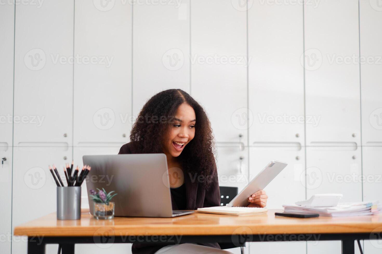 joyeux femme d'affaires est surpris comme elle lit positif nouvelles sur sa tablette à sa espace de travail, avec une portable et Stationnaire sur sa bureau. photo