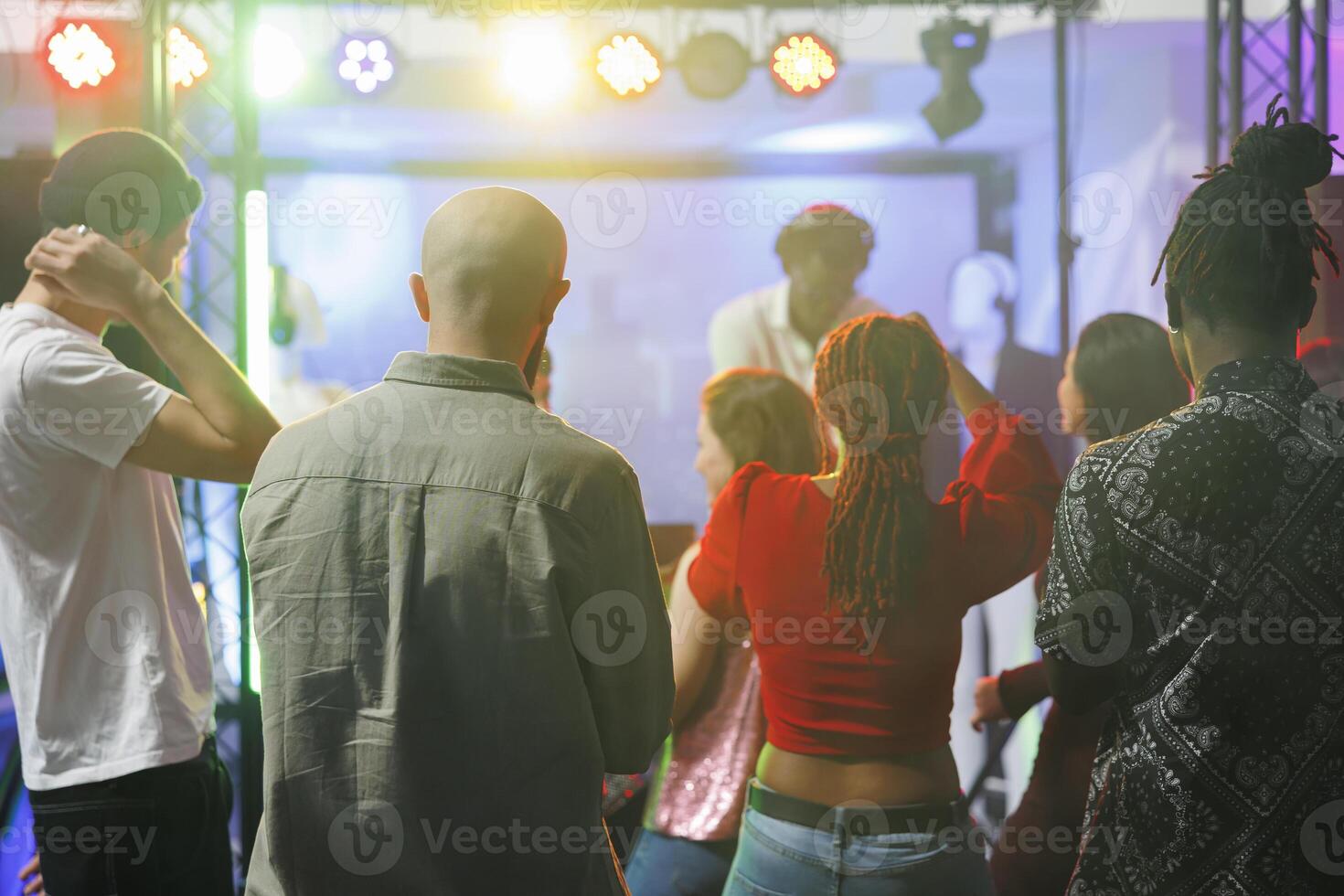 Jeune gens permanent sur piste de dance tandis que assister électronique la musique concert dans boîte de nuit. diverse Hommes et femmes foule dansant tandis que dj performant sur étape avec coloré projecteurs photo