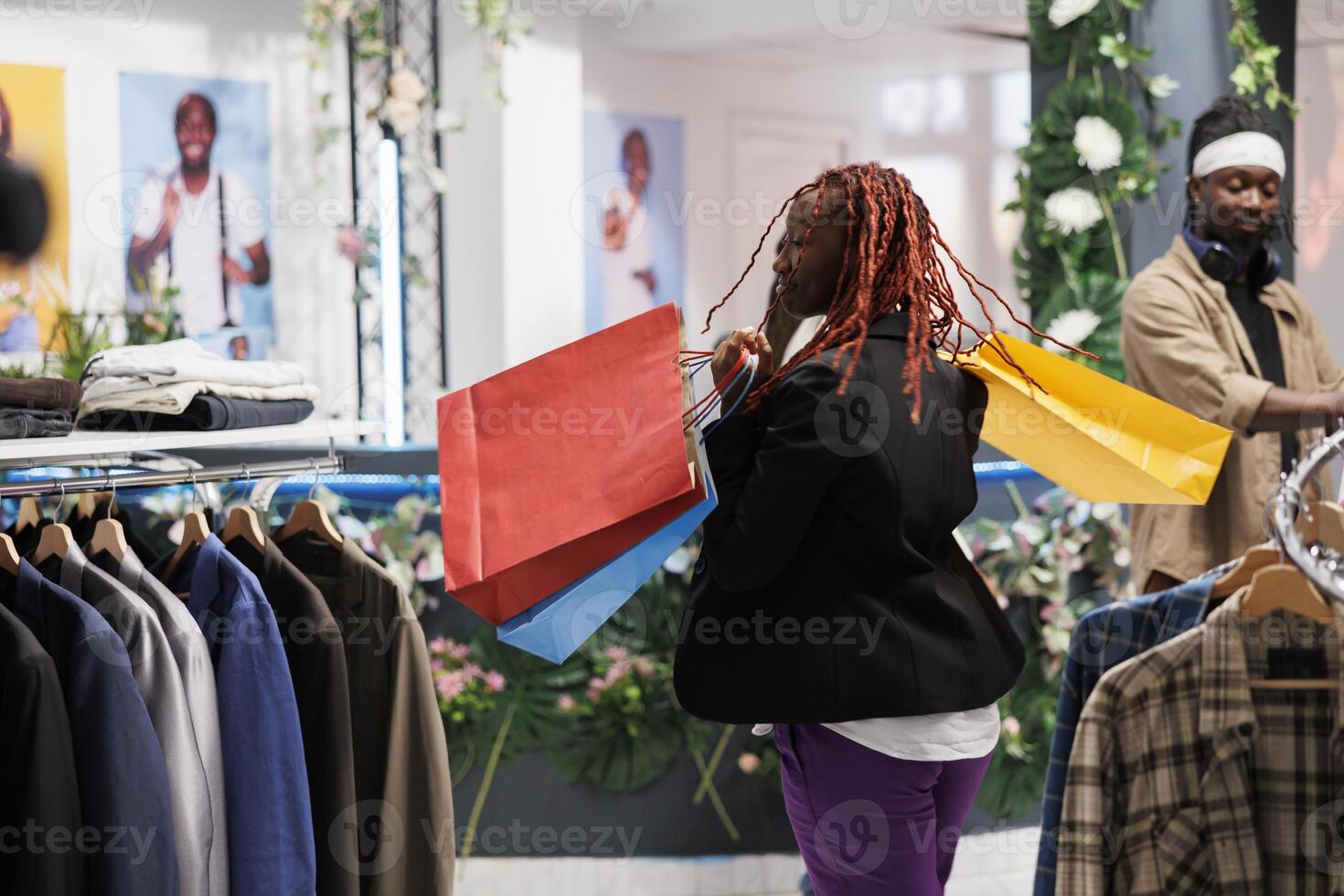 africain américain femme dansant avec joie tandis que en portant achats Sacs dans salle d'exposition. content Jeune client porter plusieurs papier paquets rempli avec réduit achats dans Vêtements boutique photo