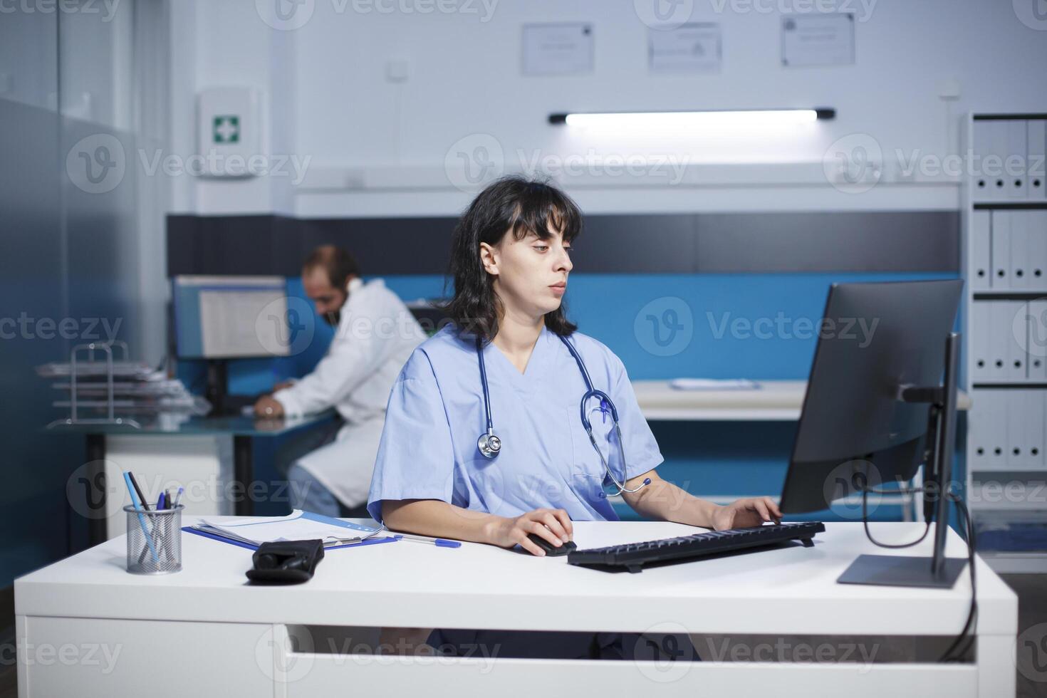 cucasien praticien est dactylographie une médical rapport à une bureau dans une hôpital Bureau en retard à nuit. femelle infirmière en train de préparer médical se soucier tandis que évaluer patient maladies symptômes. photo