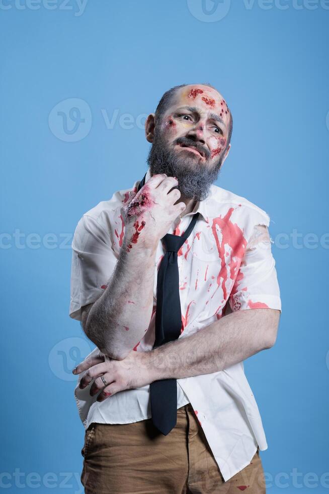 portrait de homme habillé comme zombi grimacer de douleur pour Halloween événement, portant horreur se maquiller. la personne costumé comme infecté malade créature couvert dans du sang et blessures, studio Contexte photo