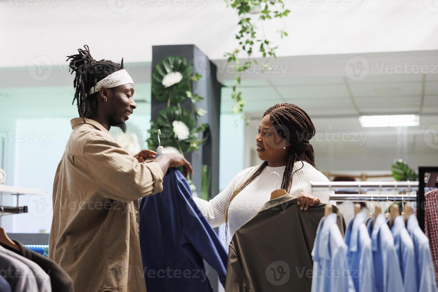 africain américain couple achat vêtements dans boutique et choisir formel veste. petite amie donnant copain tenues sur cintre à essayer et décider entre deux articles dans achats centre commercial photo