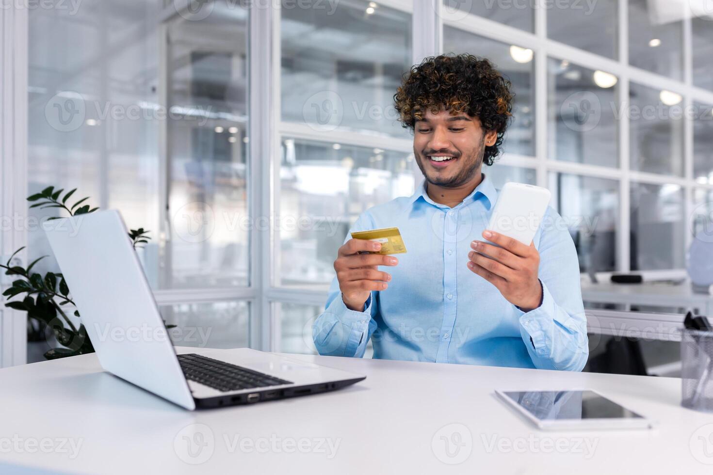 une Jeune Masculin musulman étudiant est assis dans le Bureau à une bureau avec une portable et les usages une crédit carte. détient le téléphone dans le sien mains, paie, fait du en ligne achats. photo