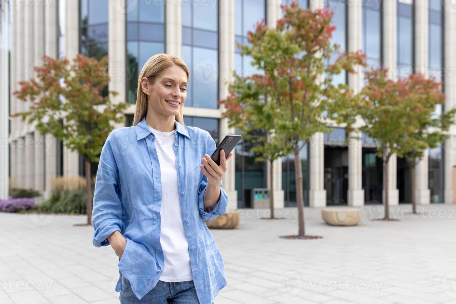 mature femme avec téléphone dans mains en marchant dans le ville, une femme d'affaires dans bleu chemise en portant téléphone intelligent dans mains, en train de lire en ligne social les réseaux, blond souriant la satisfaction est en utilisant un application. photo