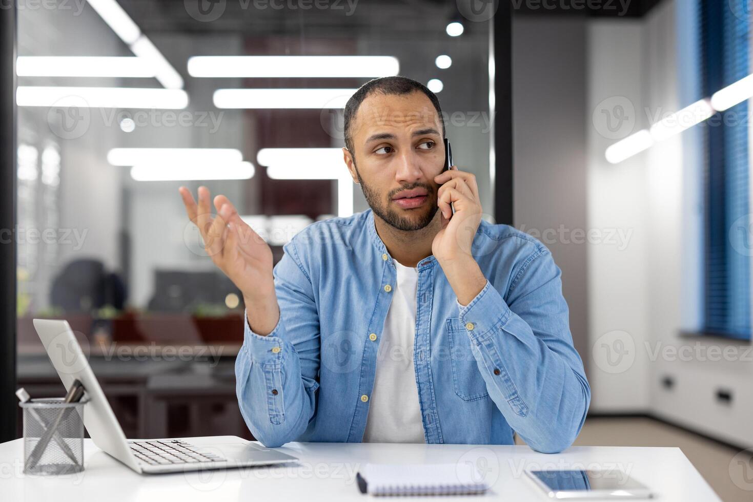 une homme est parlant sur le sien cellule téléphone tandis que séance à une bureau. il est portant une bleu chemise et il est dans une sérieux ambiance photo