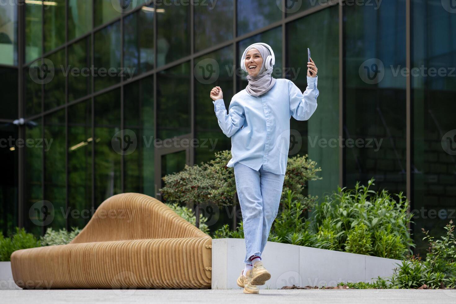 Jeune musulman femelle étudiant à l'extérieur Université Campus, content dansant à l'extérieur bâtiment, femme dans hijab écoute à la musique en utilisant téléphone et écouteurs, content souriant. photo