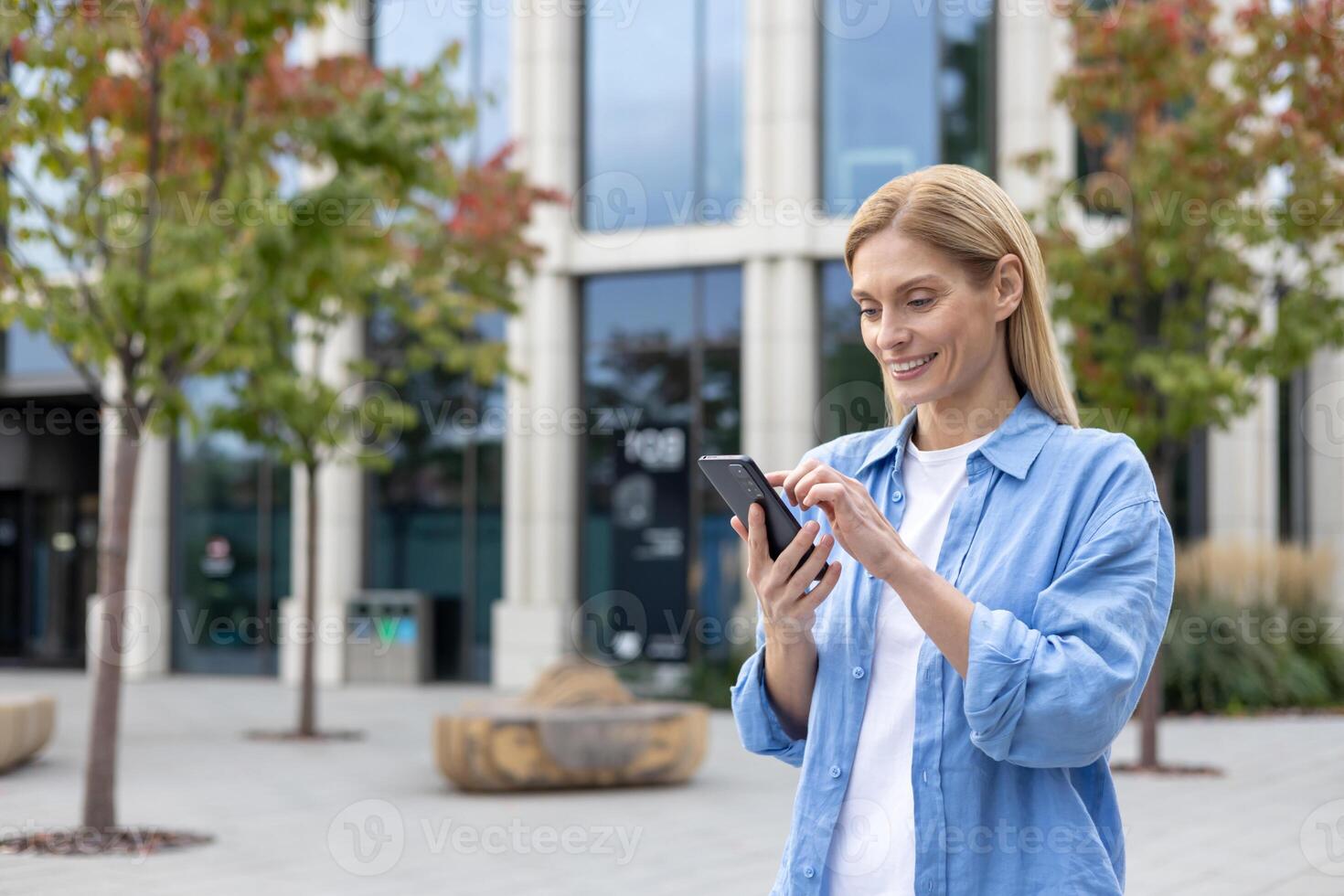 mature femme avec téléphone dans mains en marchant dans le ville, une femme d'affaires dans bleu chemise en portant téléphone intelligent dans mains, en train de lire en ligne social les réseaux, blond souriant la satisfaction est en utilisant un application. photo