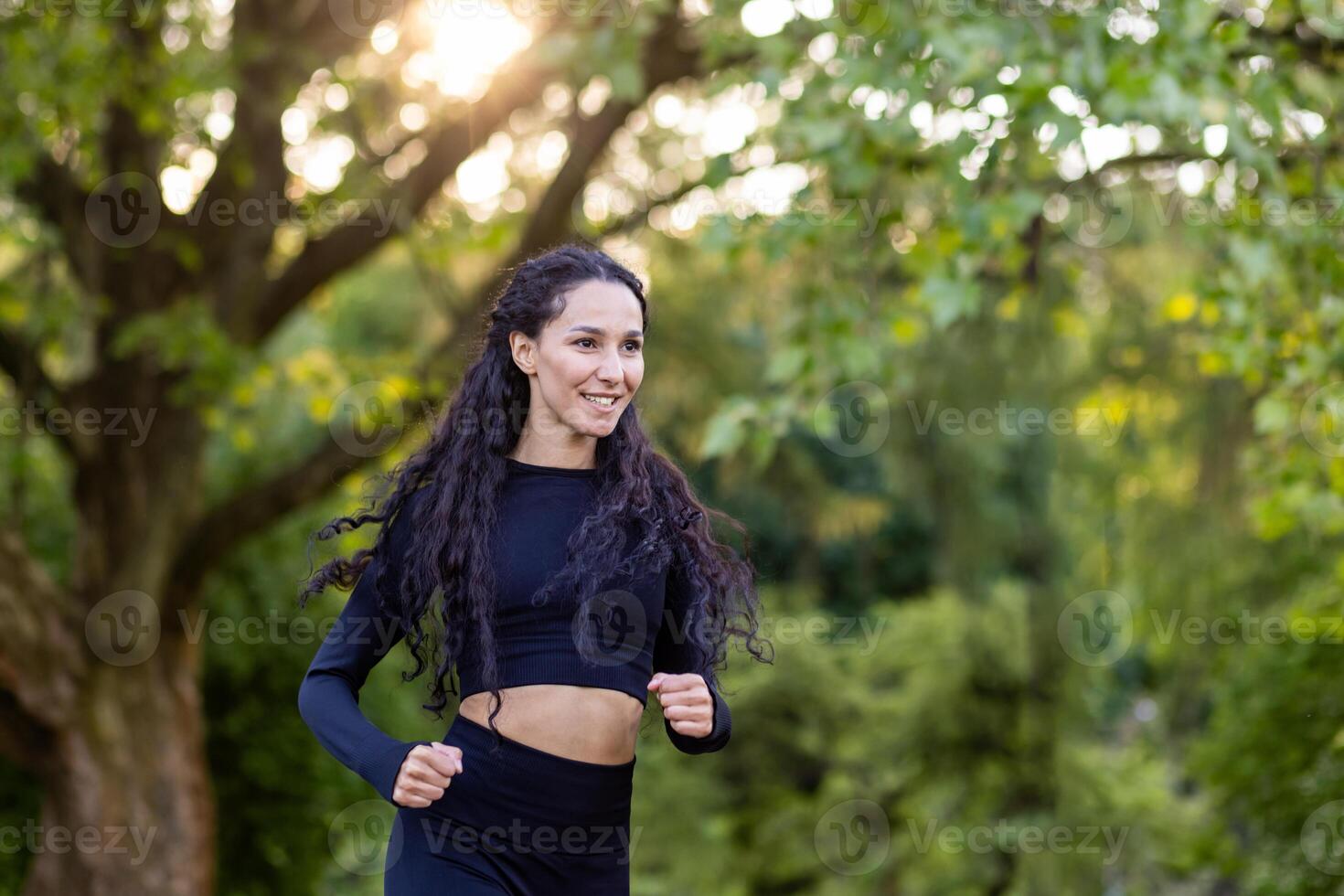 Jeune motivée et joyeux femme le jogging dans le Matin dans une Publique parc parmi des arbres, hispanique femme avec frisé cheveux, svelte dans une survêtement. photo