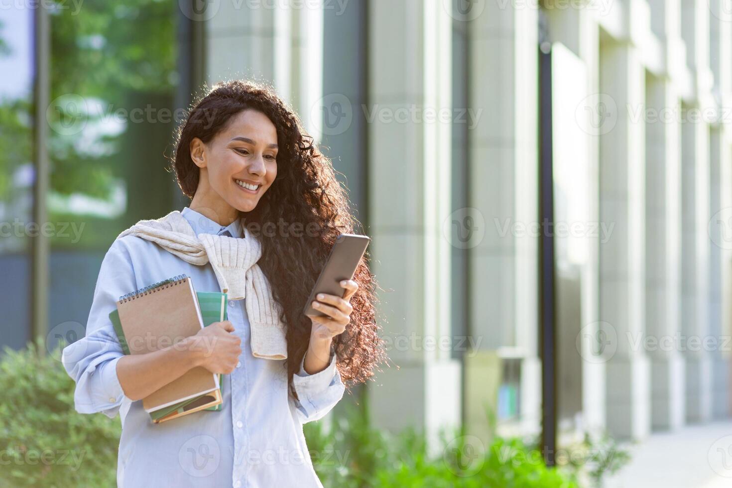 une Jeune magnifique hispanique femme des promenades ville avec une téléphone dans mains, une étudiant avec livres à l'extérieur le Campus de une Université académique bibliothèque les usages un application sur une téléphone intelligent. photo