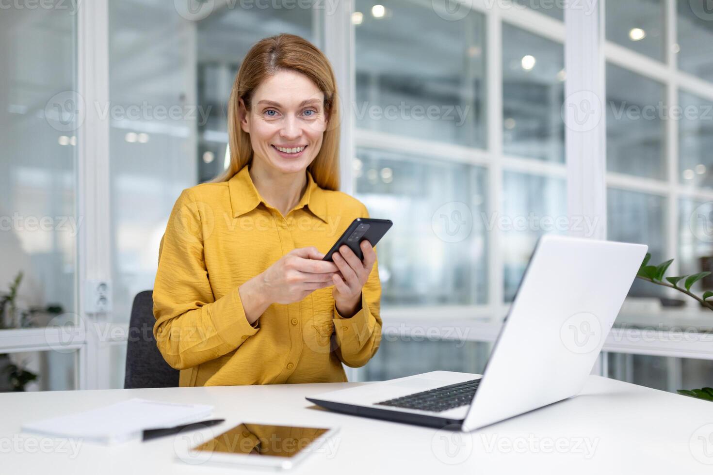portrait de une Jeune femme d'affaires travail dans le Bureau à une ordinateur portable, en portant une mobile téléphone et souriant à le caméra. photo
