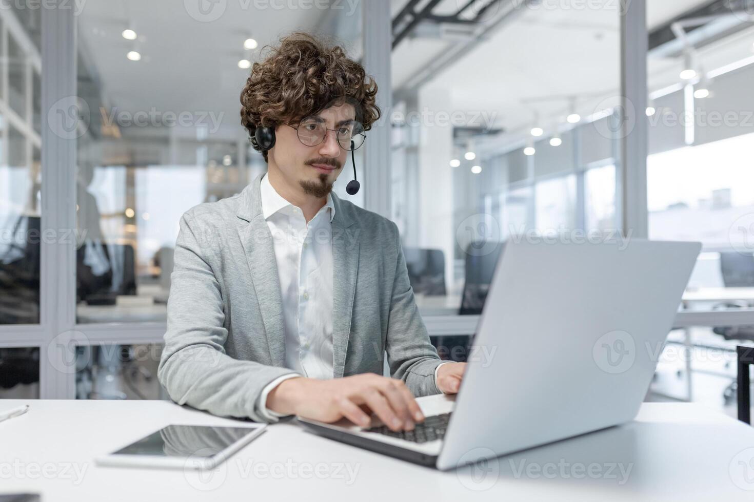 concentré Jeune adulte Masculin entrepreneur avec frisé cheveux en utilisant portable et casque dans une moderne Bureau paramètre, incorporant professionnalisme et dévouement. photo