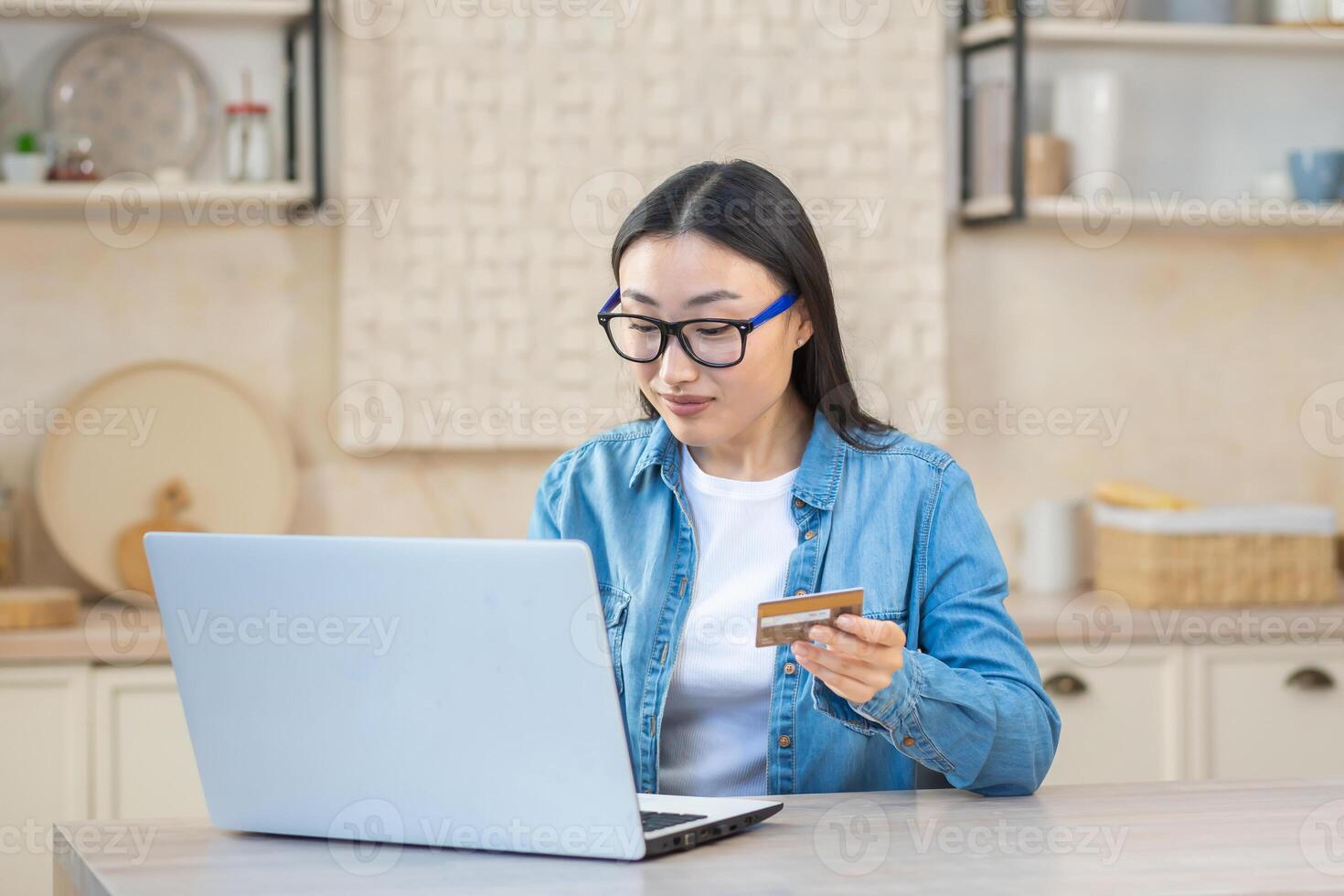 Jeune magnifique asiatique femme dans des lunettes et une denim chemise séance à le table avec une portable à maison. en portant une crédit carte, payant factures en ligne. photo