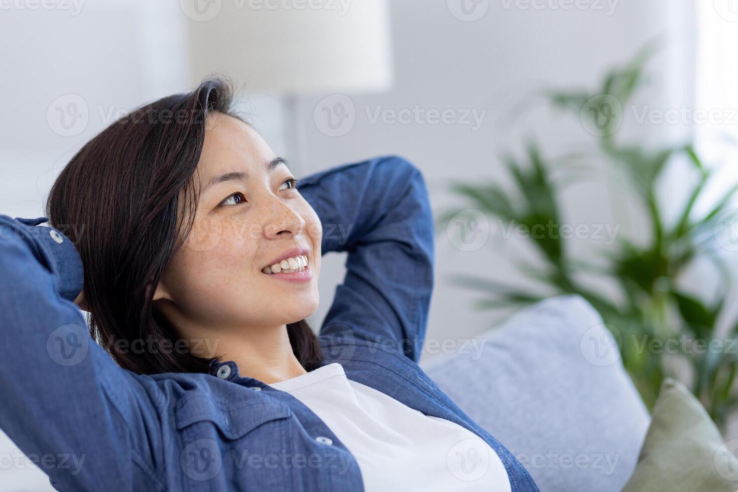 fermer photo de une Jeune magnifique asiatique à Accueil dans le vivant chambre, femme a sa mains derrière sa tête et est repos, respiration Frais air, souriant et à la recherche fenêtre, rêver content futur