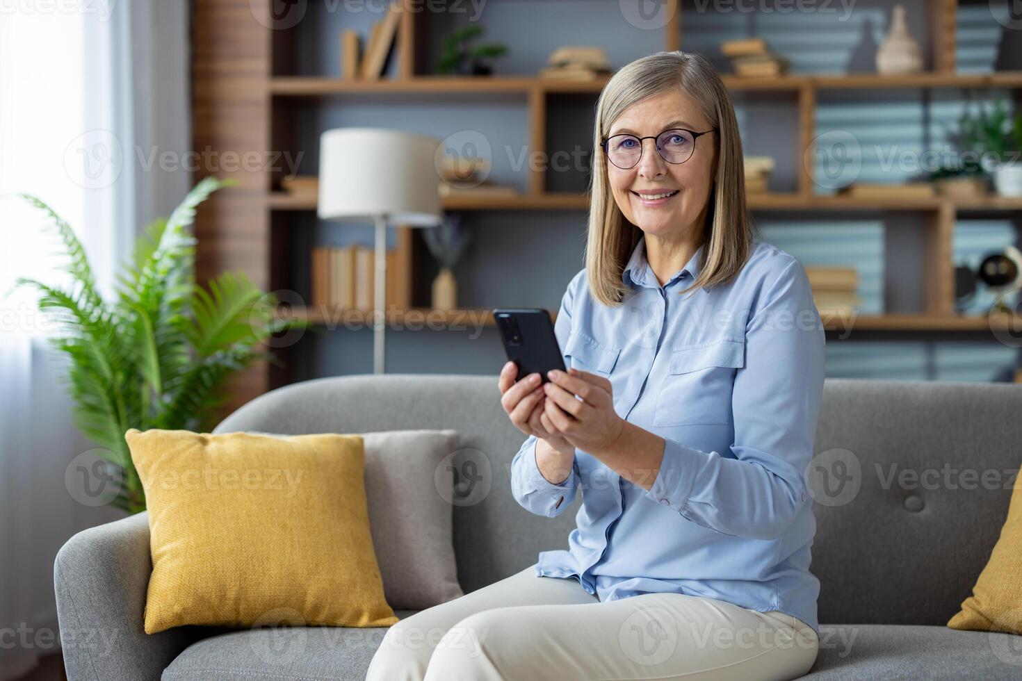 mature adulte femme confortablement assise sur une canapé, navigation sa téléphone intelligent dans une moderne vivant pièce paramètre. photo