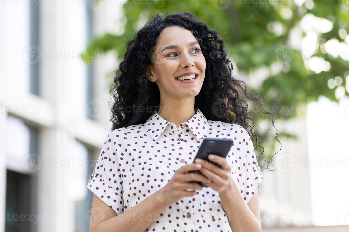 une Jeune magnifique Latin américain femme avec frisé cheveux des promenades par le ville avec une téléphone dans sa mains, une femme les usages un application sur une téléphone intelligent, parcourt l'Internet pages et social réseaux. photo