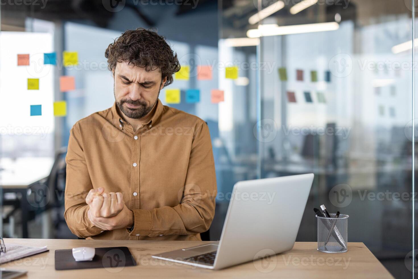 Masculin Bureau ouvrier expérience inconfort, agrippant le sien main dans douleur à le sien lieu de travail avec portable et Stationnaire autour. photo