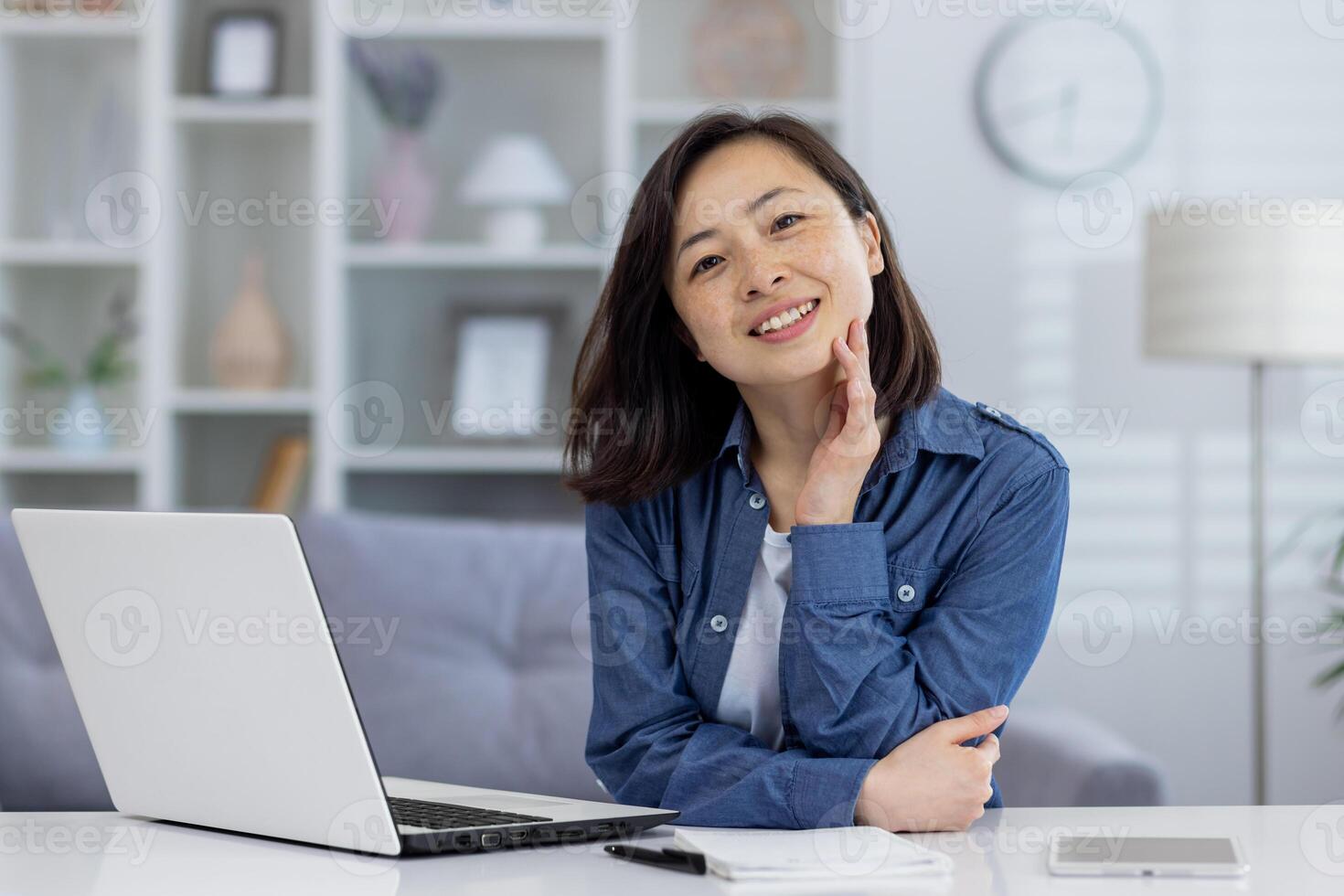 portrait de Jeune magnifique asiatique femme à maison, femme souriant et à la recherche à caméra, travail à distance dans Accueil Bureau en utilisant portable tandis que séance à table dans vivant chambre. photo