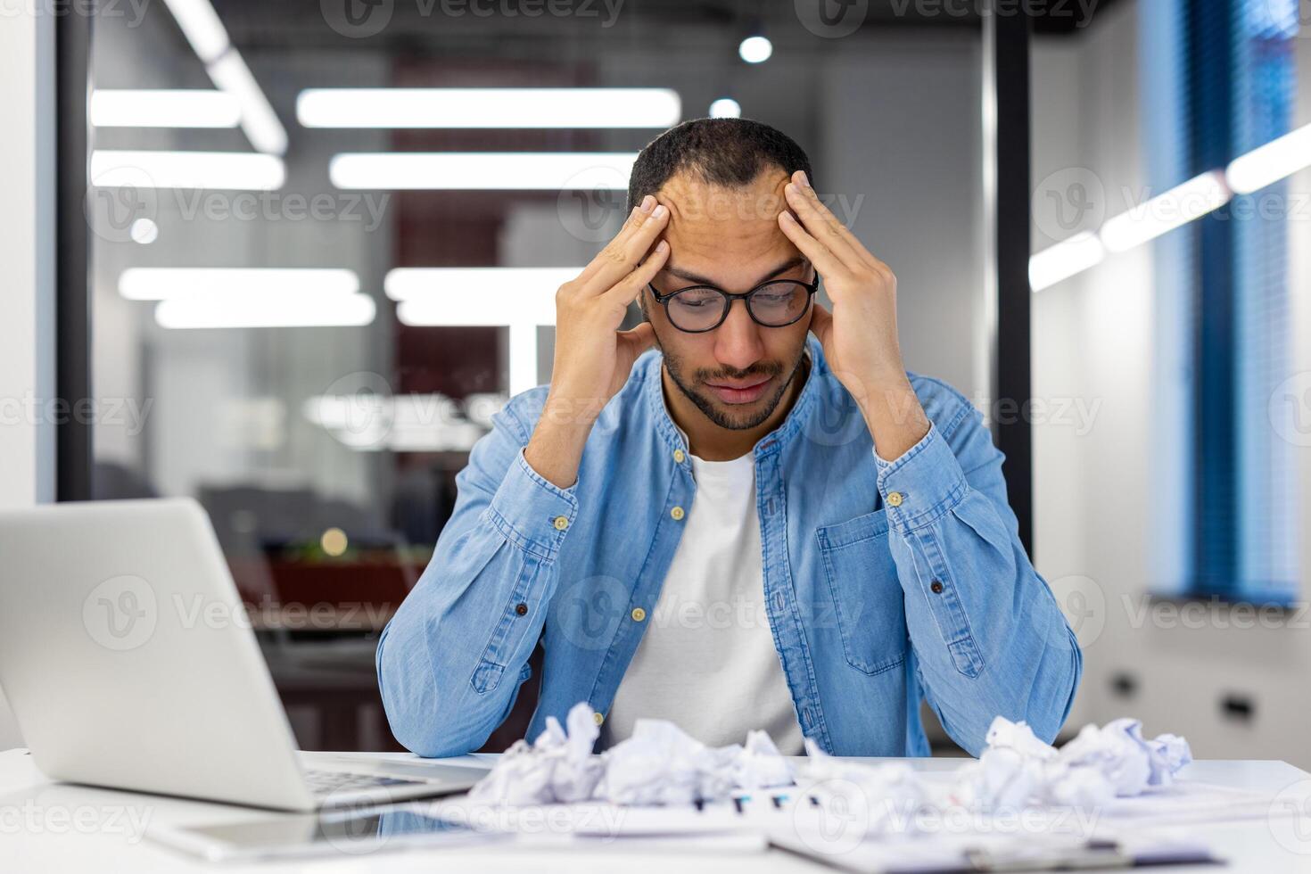 Jeune hispanique homme séance dérangé à le bureau dans le bureau, en portant le sien tête dans frustration, froissé les documents et papier. photo