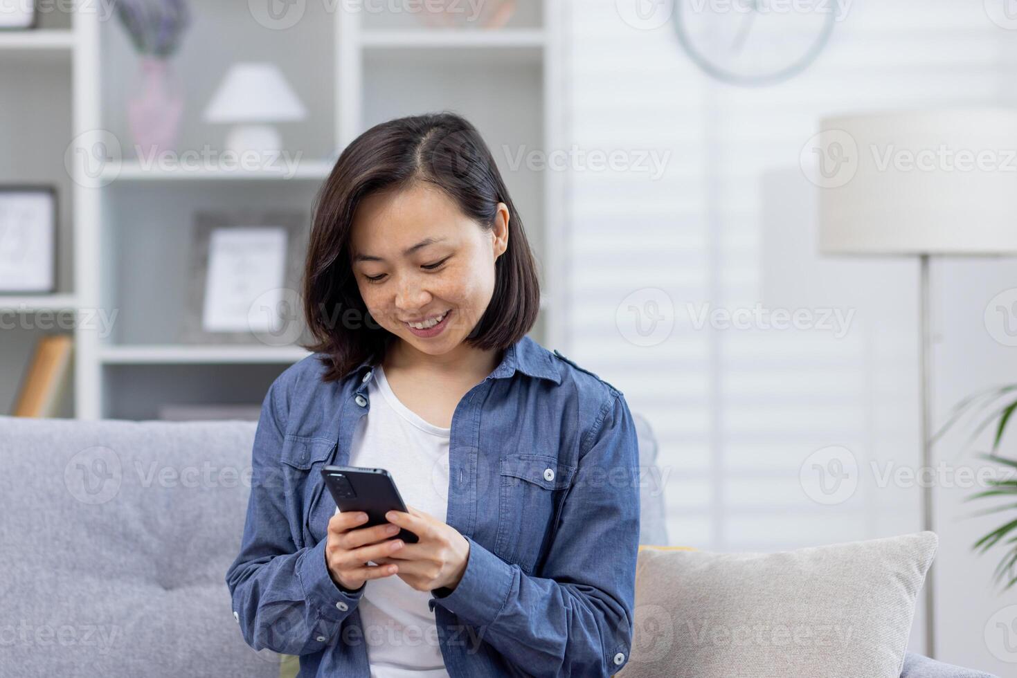 une Jeune magnifique asiatique est assis seul à Accueil sur le canapé, le femme les usages un application sur le téléphone, détient une téléphone intelligent dans sa mains, sourit Heureusement, les usages le application, parcourt en ligne pages. photo