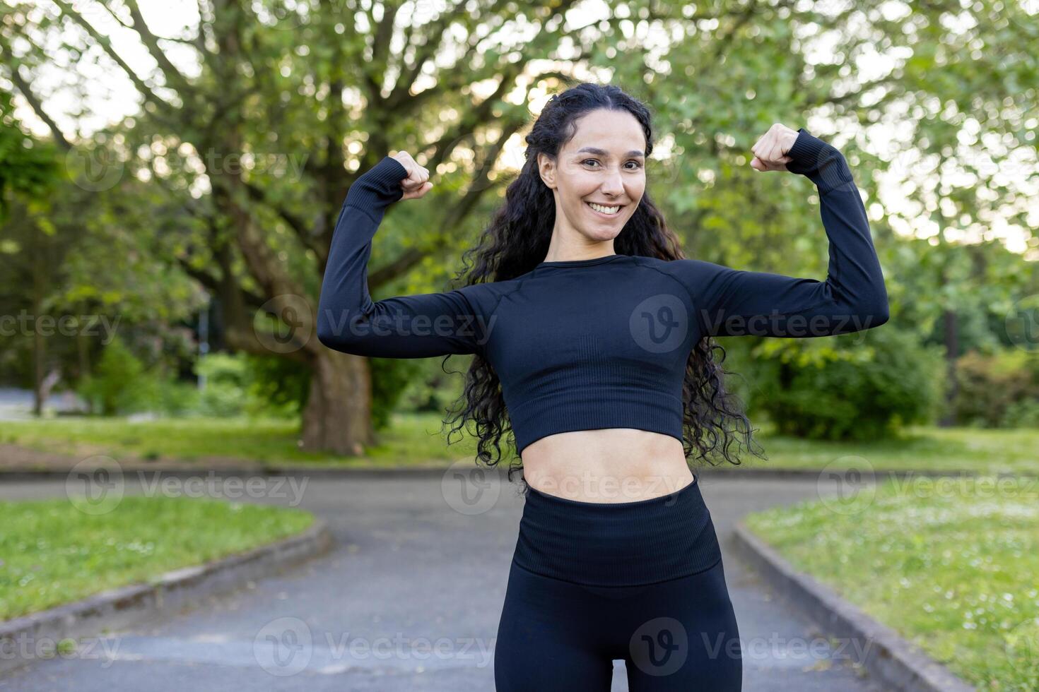 une de bonne humeur hispanique femme fléchit sa muscles en toute confiance dans une parc, affichage force et aptitude dans une Naturel paramètre. habillé dans sportif noir tenue, elle sourit largement. photo