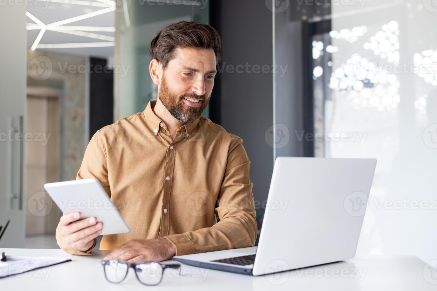 une Jeune Masculin programmeur et développeur est assis dans le Bureau à le table et travaux sur une portable et avec une tablette. pourparlers sur une appel. photo