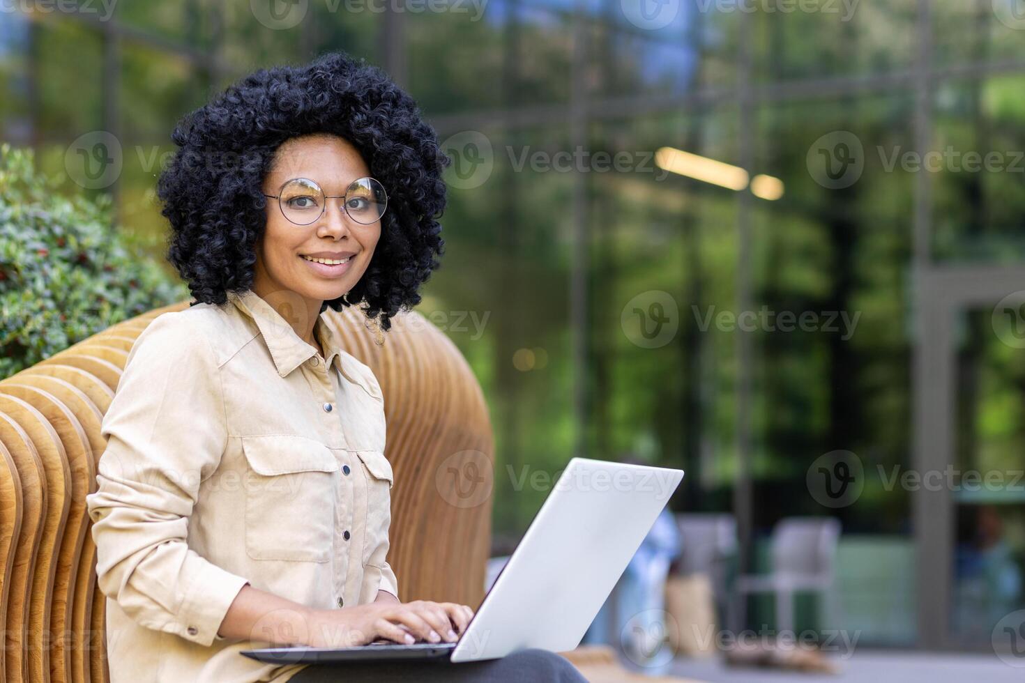 portrait de un africain américain Jeune femelle étudiant séance sur une banc à l'extérieur une Université Campus en train d'étudier en ligne sur une portable. souriant et à la recherche à le caméra. photo