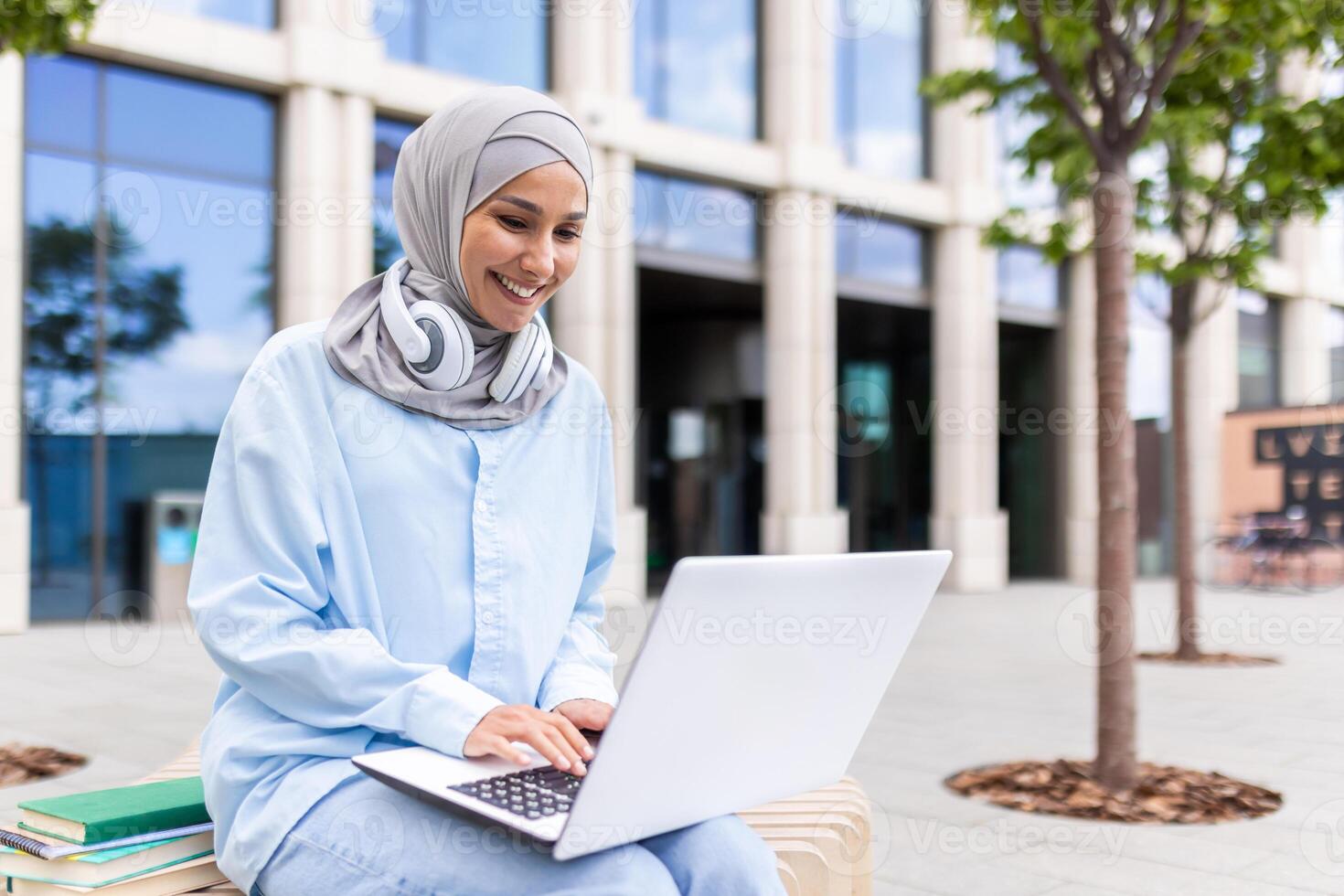 Jeune musulman femme portant une hijab travail sur une portable tandis que séance sur une banc à l'extérieur Université bâtiments. photo