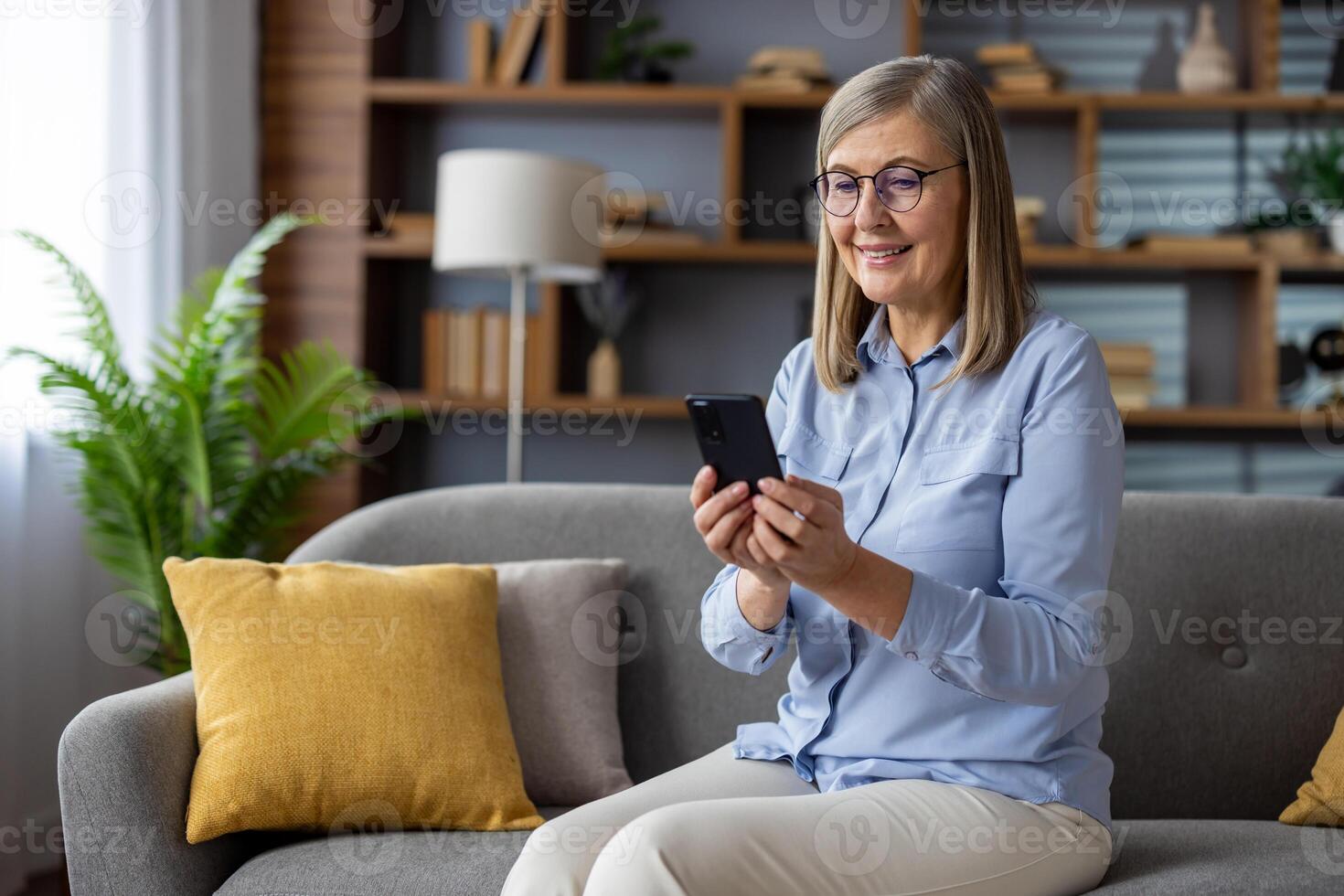 Sénior aux cheveux gris femme séance joyeusement sur canapé à maison, retraité en portant téléphone dans mains, en utilisant app sur téléphone intelligent, dactylographie texte message, appel, et navigation social réseaux avec une sourire. photo