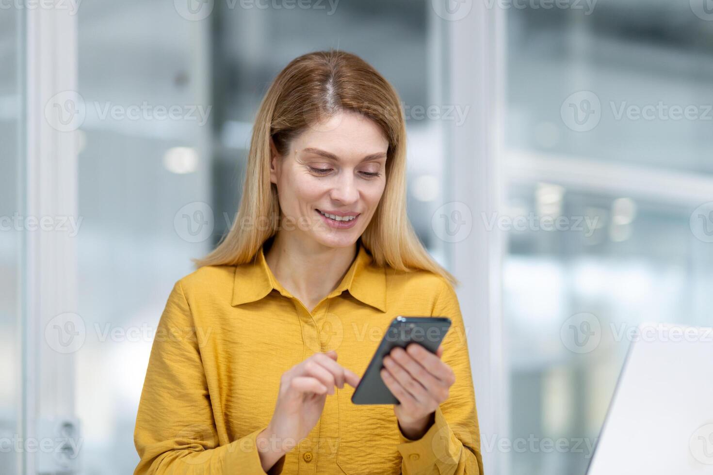 mature expérimenté affaires femme à l'intérieur Bureau à lieu de travail, employé séance à bureau souriant en portant téléphone dans main, patron en utilisant application sur téléphone intelligent, navigation l'Internet pages. photo