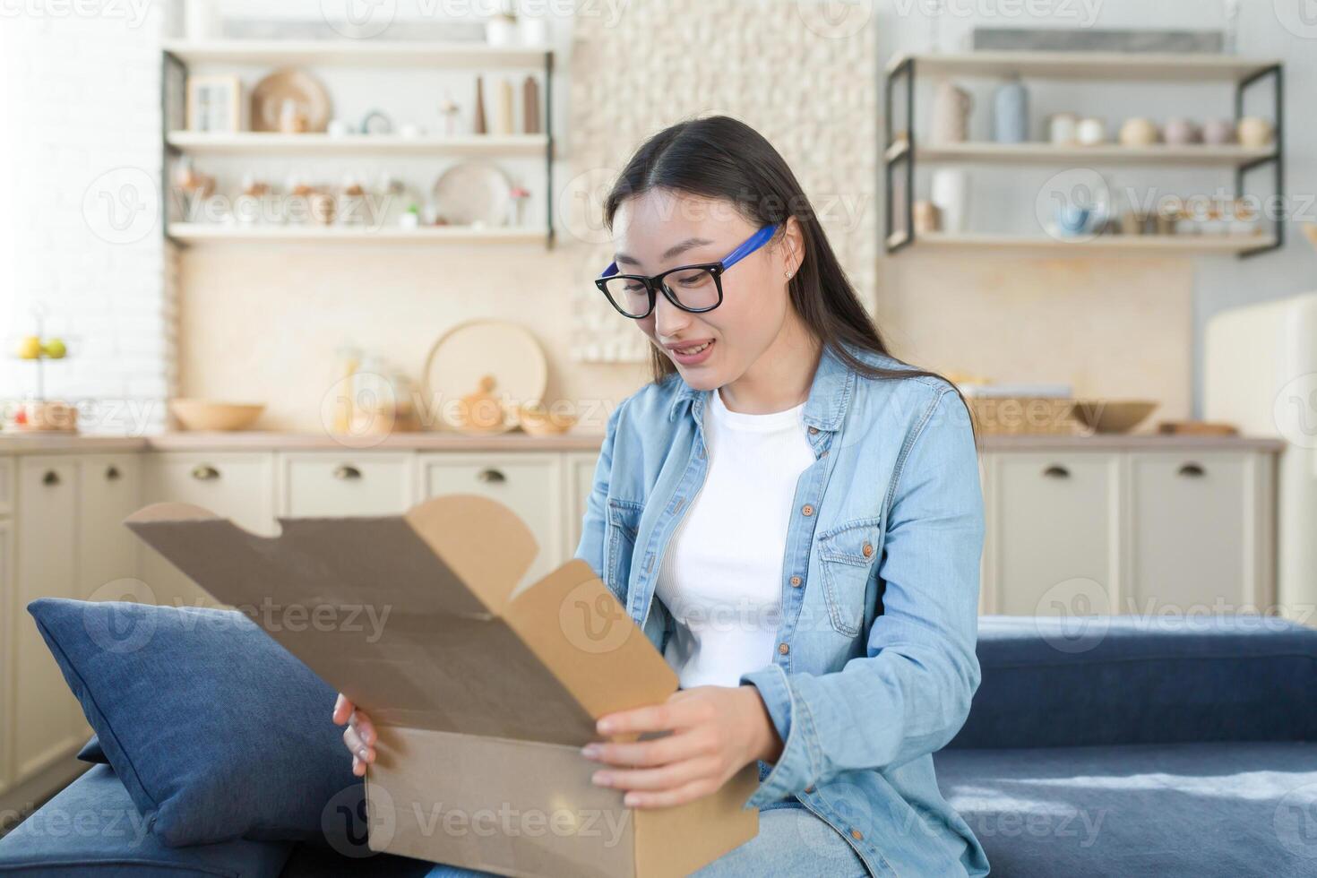 une Jeune magnifique asiatique femme reçu une paquet par une courrier avec Accueil livraison. séance sur le canapé à Accueil et ouverture le boîte, en ligne commande de le magasin. photo