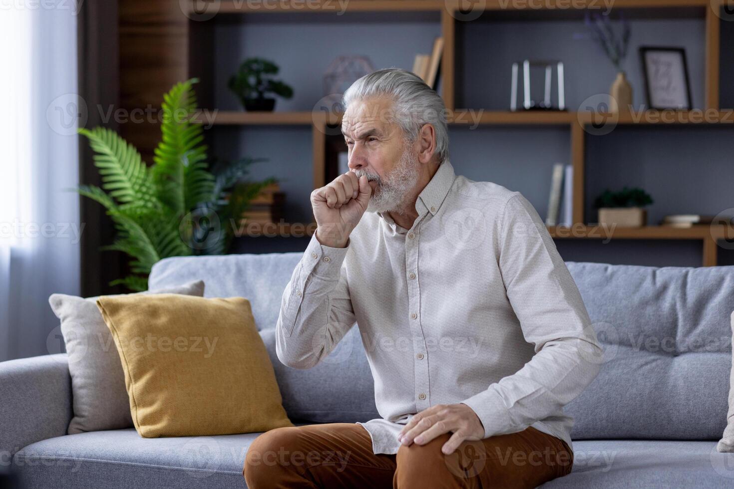 mature homme sentiment indisposé, tousser tandis que séance seul sur une canapé dans une bien décoré, contemporain vivant chambre. concept de vieillissement et santé questions. photo