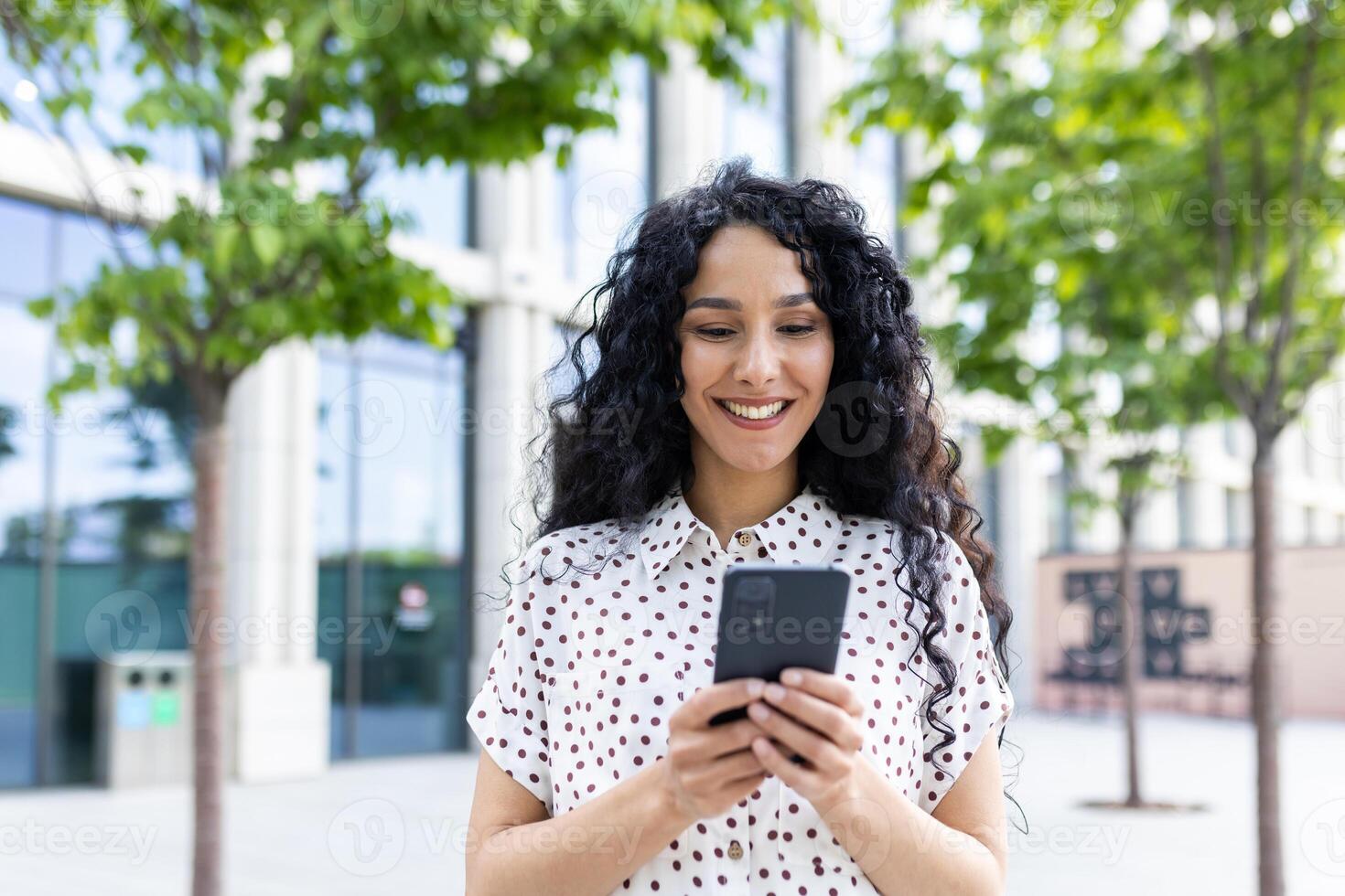 une Jeune magnifique femme des promenades par le ville avec une téléphone dans sa mains, sourit avec contentement, les usages une téléphone intelligent application, parcourt l'Internet pages, les types une texte message, social réseaux. photo