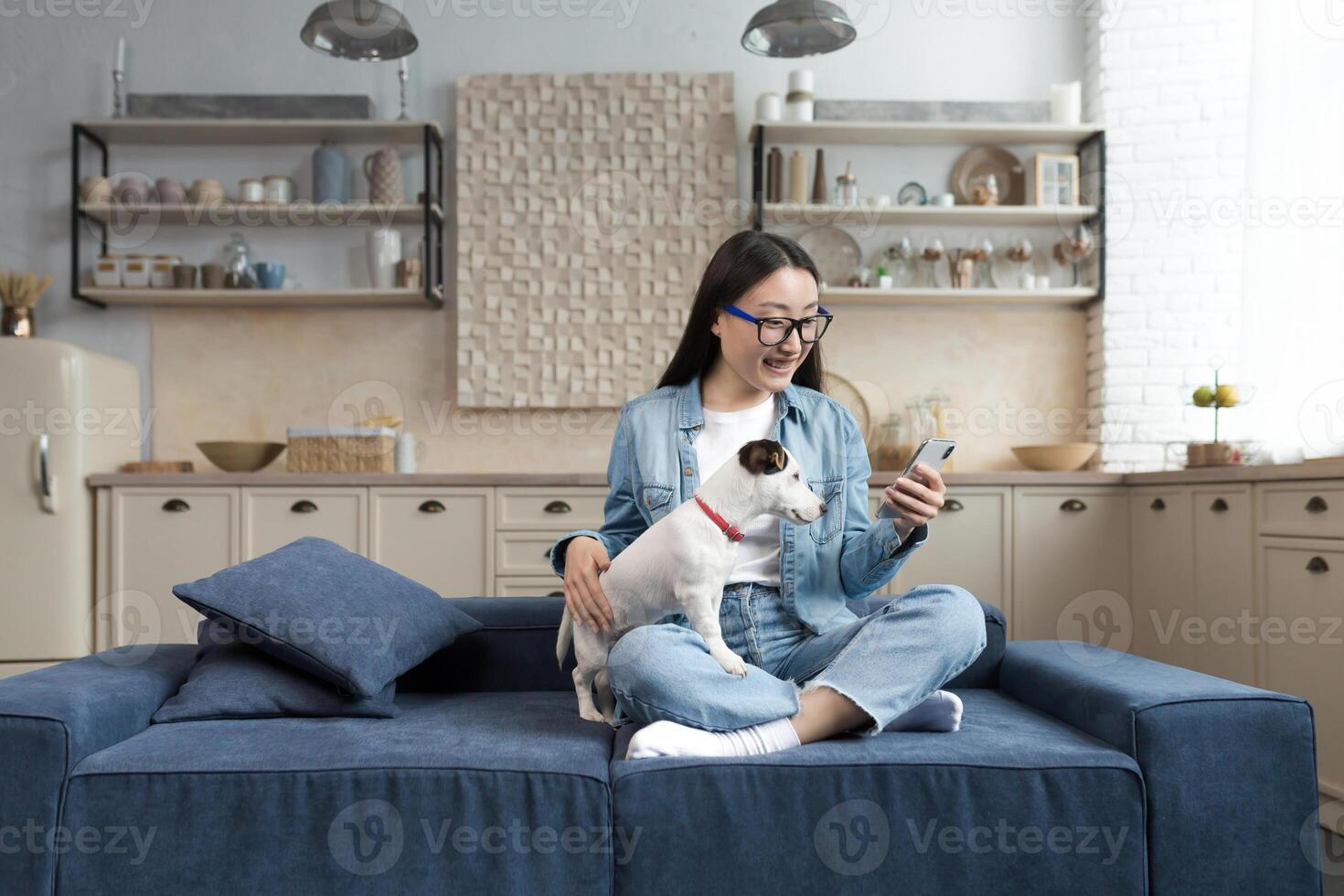 Jeune magnifique asiatique femme avec animal de compagnie jack Russell terrier parlant sur appel avec amis, femme à Accueil dans cuisine en utilisant téléphone intelligent pour éloigné communication. photo