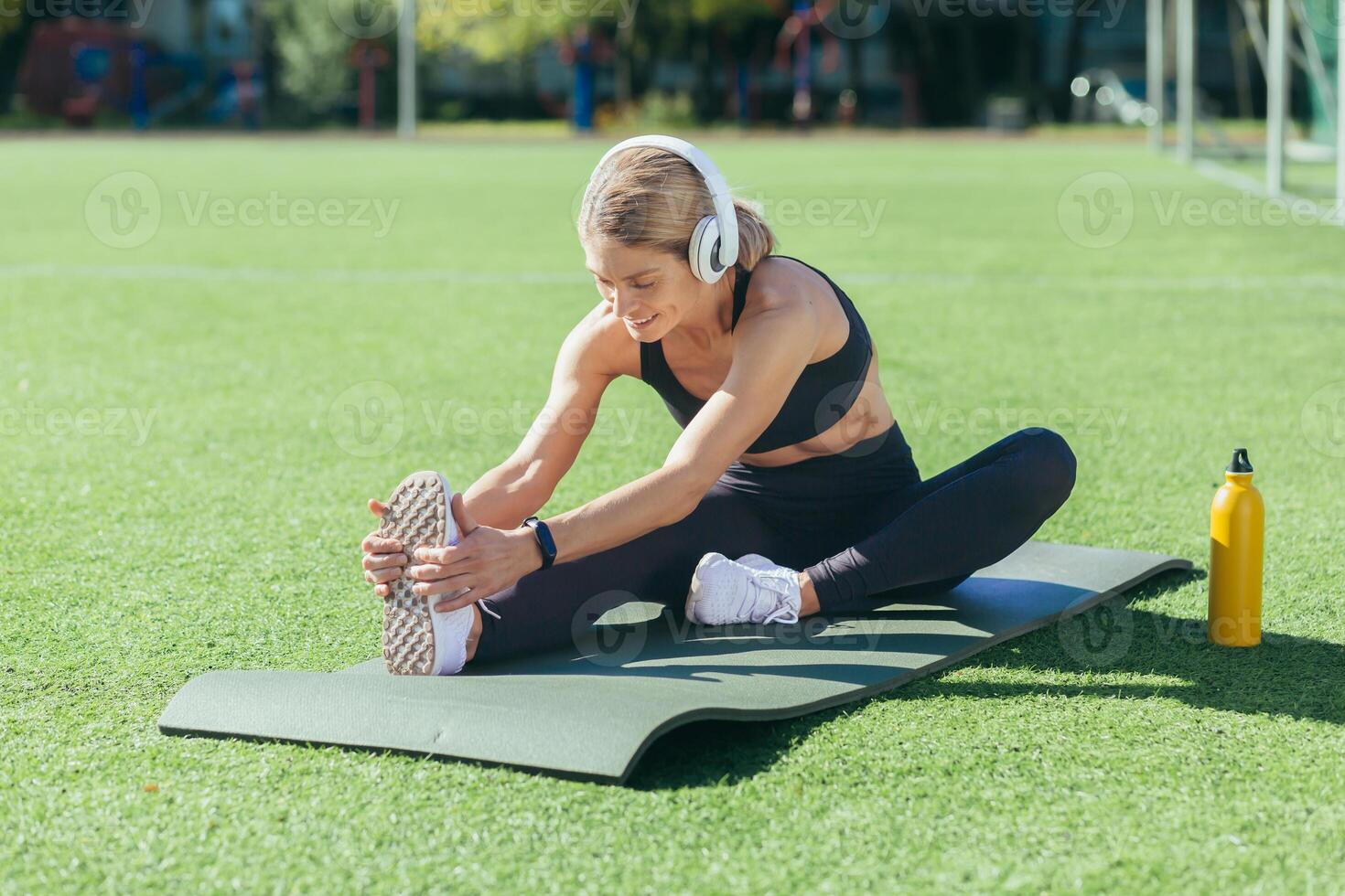 blond femme Faire physique des exercices séance sur des sports tapis, sportive élongation muscles, écoute à la musique dans écouteurs et souriant, Matin stade sur ensoleillé journée. photo